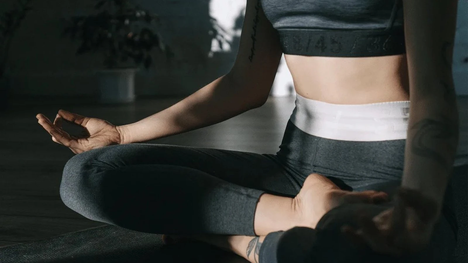 A woman in gray athletic wear sitting cross-legged on a studio floor in a meditation pose.